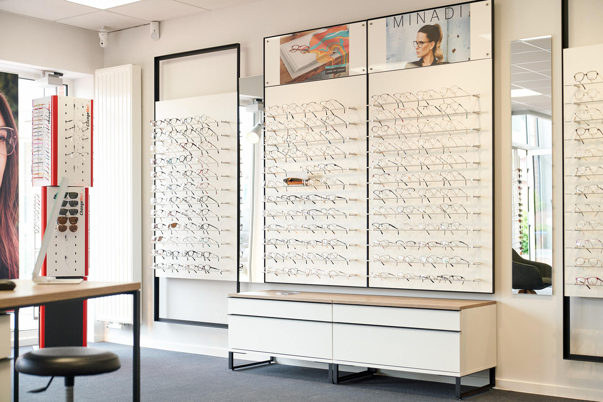 Elegante Brillenwand in einem Optikergeschäft mit einer großen Auswahl an modernen Fassungen, stilvollen Spiegeln und einer strukturierten Präsentation.